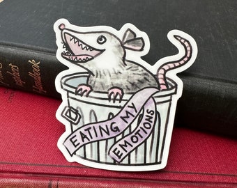May include: A sticker featuring a cartoon opossum with a toothy grin, sitting in a trash can. The opossum is wearing a purple ribbon that says "Eating My Emotions".