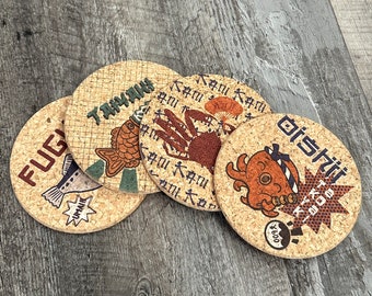 May include: Four round cork coasters with Japanese text and images of fish, crabs, and other sea creatures. The coasters are brown with white and black accents.