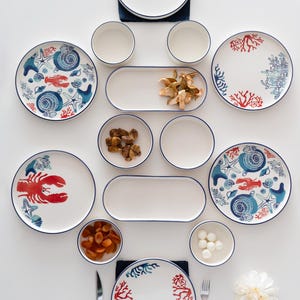 Pode incluir: Conjunto de louças de cerâmica branca com tema náutico. Inclui pratos, tigelas e travessas decoradas com lagostas vermelhas, corais e conchas. Cada peça tem uma borda azul.