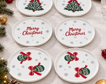 以下が含まれることがあります： クリスマスデザインの白い円形セラミック製コースター6枚セット。2枚には赤いリボンの付いた緑のクリスマスツリー、2枚には赤い文字でMerry Christmas、残り2枚には赤いリボンの付いた緑のオーナメントが描かれています。