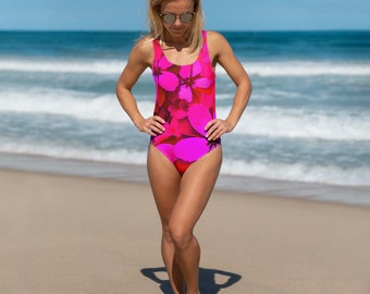 Op de afbeelding: Een badpak met een levendig bloemenpatroon in roze, magenta en rode tinten. Het badpak heeft een ronde hals en een klassiek silhouet. De achtergrond toont een zandstrand en de oceaan.