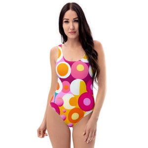 Peut inclure: Un maillot de bain une pièce avec un motif rétro vibrant. Le maillot de bain présente un motif de cercles superposés dans des tons d'orange, de rose, de jaune et de blanc sur un fond magenta foncé. Le décolleté et le dos sont visibles.