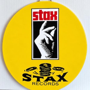 May include: A circular yellow wall sign featuring the Stax logo in red and black. The design includes a black and white graphic of a hand snapping fingers inside a rectangular border, with a stack of vinyl records and the text Stax Records below.