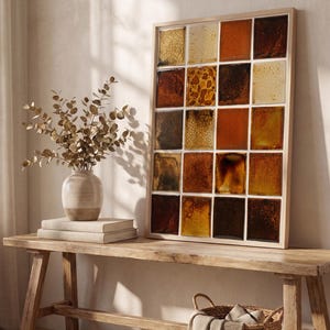 Op de afbeelding: Een rechthoekig wandkunstwerk met een raster van twintig vierkante keramische tegels in diverse tinten amber, bruin en oker. Elke tegel heeft een unieke glanzende glazuurafwerking met gebarsten of gevlekte texturen, gevat in een lichthouten lijst.