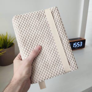Pode incluir: Um caderno retangular com uma capa de tecido texturizado na cor creme, com um padrão em relevo tipo pipoca. O caderno possui uma faixa elástica vertical plana, em bege claro, que envolve a capa frontal e uma pequena alça correspondente na base.