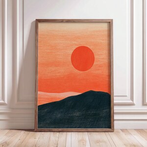 Op de afbeelding: Een ingelijste kunstdruk met een landschapsillustratie met een grote, effen oranje cirkelvormige zon in een oranje gradiëntlucht boven een donker, getextureerd houtskoolkleurig gebergte met zichtbare potloodachtige streken.