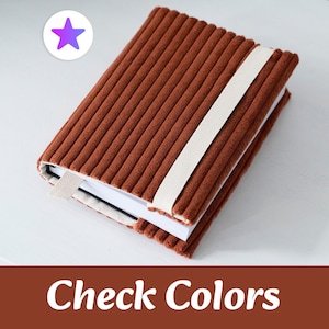 Pode incluir: Um caderno retangular com uma capa de tecido de veludo cotelê texturizado na cor ferrugem. O caderno é preso por uma faixa elástica larga na cor creme que envolve o exterior. Uma pequena aba de marcador de página em tecido creme sobressai da lateral.