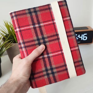 Pode incluir: Um caderno retangular com uma capa de tecido em xadrez vermelho, preto e branco. O caderno é preso por uma faixa elástica larga na cor creme que envolve verticalmente a capa frontal e inclui um marcador de página de tecido a condizer.