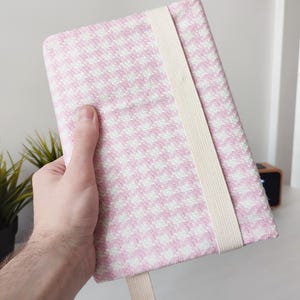 Pode incluir: Uma capa retangular para caderno com um padrão tecido de pé-de-galinha em rosa e branco. A capa é fixada com uma faixa elástica vertical larga na cor creme que envolve a parte frontal, com uma pequena alça elástica a condizer na borda inferior.