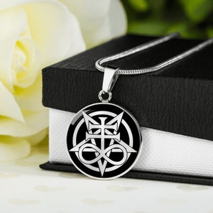 Op de afbeelding: Een zilveren ketting met een zwarte hanger met een wit symbool van een kruis met een oneindigheidssymbool en een pentagram. De hanger ligt op een zwarte doos met een witte binnenkant.