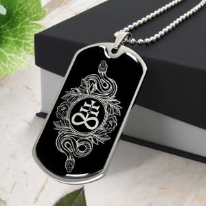 Op de afbeelding: Een zwarte hondenpenning met een zilveren ketting. De hondenpenning heeft een wit ontwerp van een slang die om een cirkel met een symbool erin gewikkeld is. Het symbool is een kruis met een cirkel eromheen en het getal 8 in de cirkel.