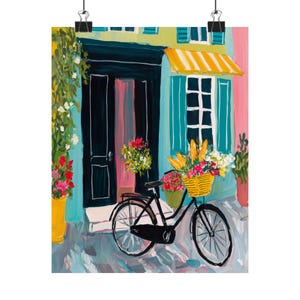 May include: An artistic print shows a European street scene. A black bicycle with a yellow basket of flowers and baguettes is parked in front of a building with a teal door and shutters, and a yellow awning. Pink and yellow flowers are in pots.