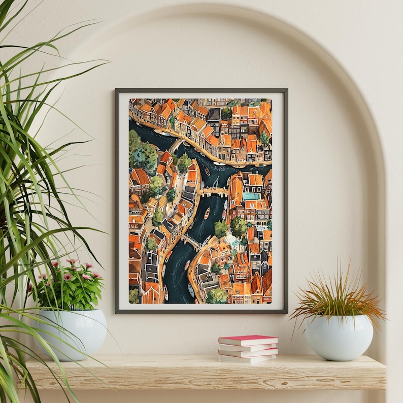 May include: A watercolour painting of a city with canals and bridges. The buildings are painted in shades of orange, yellow, and red. The canals are a deep blue colour. The painting is framed in a grey frame.