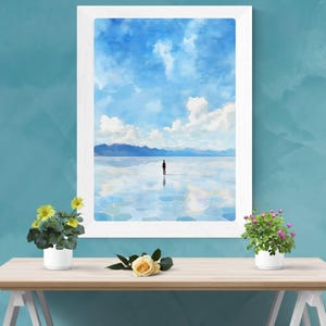 May include: A framed watercolor painting of a lone figure standing on a reflective surface with a mountain range in the background and a blue sky with white clouds.