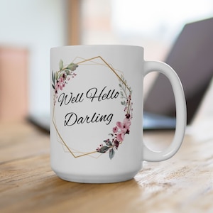 May include: White ceramic mug with a gold geometric frame and floral design. The text "Well Hello Darling" is printed in black inside the frame.