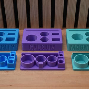 Op de afbeelding: Drie 3D-geprinte organizers in blauw, paars en teal, elk gelabeld met een chemische naam: ALKALINITEIT, CALCIUM en MAGNESIUM. Elke organizer heeft verschillende cirkelvormige en rechthoekige compartimenten.