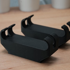 May include: Two black, 3D-printed cradles, each with a rectangular base and curved arms. The cradles are designed to hold an item. The background features a wooden surface and blurred white objects.