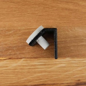 Op de afbeelding: Een zwarte kunststof hoekbeugel met een witte kunststof stop. De beugel is ontworpen om te worden gebruikt voor het bevestigen van een plank of ander object aan een muur.