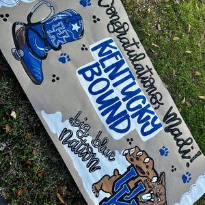 May include: A hand-painted banner with a brown background and white and blue accents. The banner features a blue cowboy boot, paw prints, and the words "Congratulations Madi!" and "Kentucky Bound." A wildcat graphic is also included.