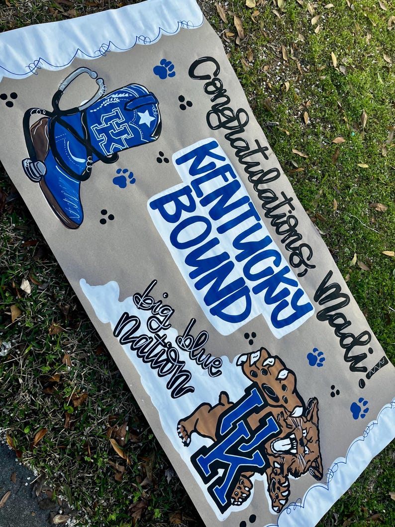 May include: A hand-painted banner with a brown background and white and blue accents. The banner features a blue cowboy boot, paw prints, and the words "Congratulations Madi!" and "Kentucky Bound." A wildcat graphic is also included.