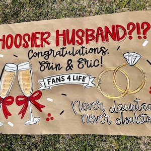 May include: A hand-painted sign on brown paper with the text "HOOSIER HUSBAND?!" in red. It includes champagne glasses, wedding rings, and the names "Erin & Eric". The sign also says "FANS 4 LIFE".