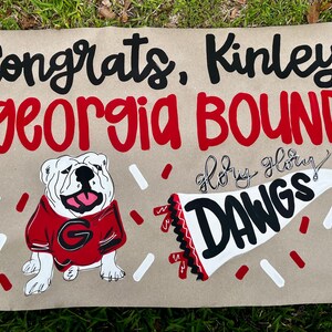 May include: A hand-painted sign on brown paper with the words "Congrats, Kinley!" and "Georgia Bound" in black and red. It features a bulldog in a red jersey and a pennant that says "DAWGS".