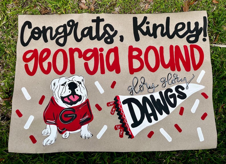 May include: A hand-painted sign on brown paper with the words "Congrats, Kinley!" and "Georgia Bound" in black and red. It features a bulldog in a red jersey and a pennant that says "DAWGS".
