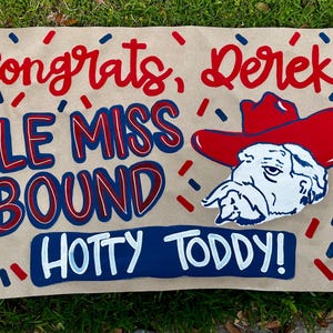 May include: A tan sign with red and blue text that reads "Congrats, Derek!" and "Ole Miss Bound." A cartoon of a bulldog wearing a red cowboy hat is also featured. The sign also says "Hotty Toddy!" in blue and white.