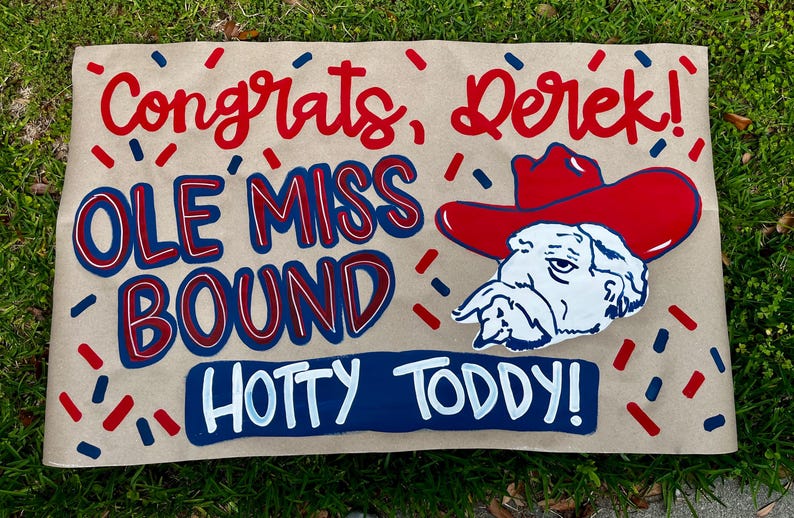 May include: A tan sign with red and blue text that reads "Congrats, Derek!" and "Ole Miss Bound." A cartoon of a bulldog wearing a red cowboy hat is also featured. The sign also says "Hotty Toddy!" in blue and white.
