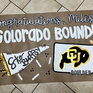 May include: A handmade sign on brown paper with the words "Congratulations, Miles! COLORADO BOUND!" in white and black lettering. A white megaphone with "Sko Buffs!" is on the left, and a black and gold buffalo logo is on the right.