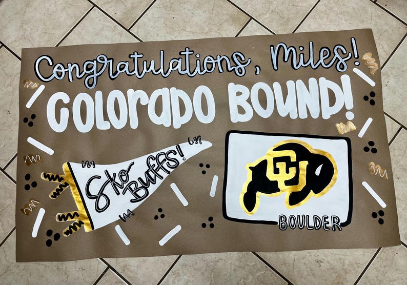 May include: A handmade sign on brown paper with the words "Congratulations, Miles! COLORADO BOUND!" in white and black lettering. A white megaphone with "Sko Buffs!" is on the left, and a black and gold buffalo logo is on the right.