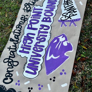 May include: A handmade banner with the words "Congratulations, Brixton! High Point University Bound!" in black and purple lettering. The banner also features a purple panther head and a white pennant with "Be Panthers" written on it.