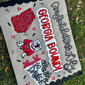May include: A hand-painted sign on tan paper with black scalloped edges. The sign features the words "GEORGIA BOUND!" in red and white, along with a bulldog graphic and the words "GO DAWGS!" in white. The sign also says "Congratulations Lily".