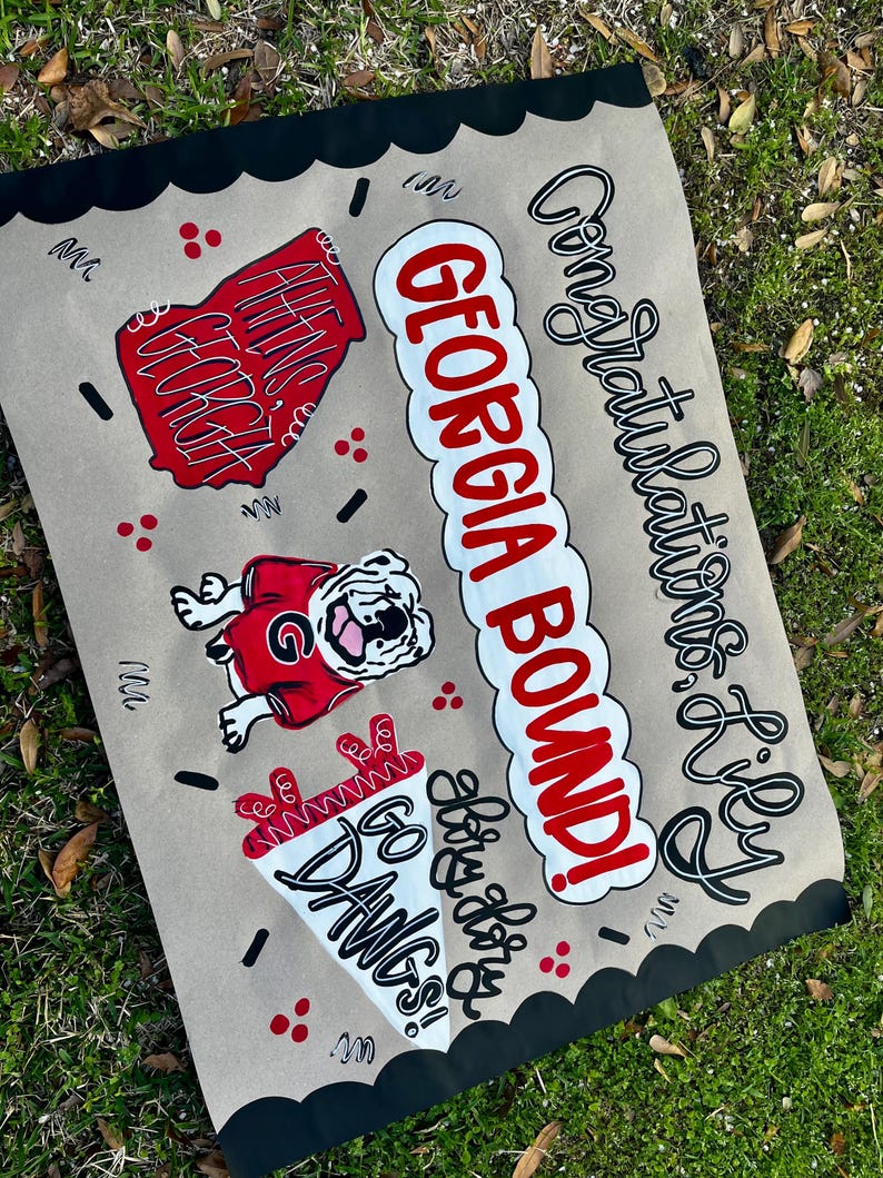 May include: A hand-painted sign on tan paper with black scalloped edges. The sign features the words "GEORGIA BOUND!" in red and white, along with a bulldog graphic and the words "GO DAWGS!" in white. The sign also says "Congratulations Lily".