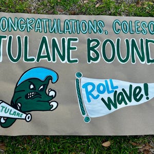 May include: A hand-painted sign on brown paper with blue borders and green text that reads "CONGRATULATIONS, COLESON! TULANE BOUND!" with a Tulane mascot and a flag that says "Roll Wave!"