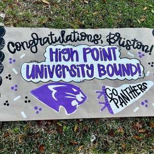 May include: A handmade banner with the text "Congratulations, Briston! High Point University Bound!" in black and purple. It features a purple panther logo and a white pennant that says "Go Panthers". The banner has black borders and is on a tan background.