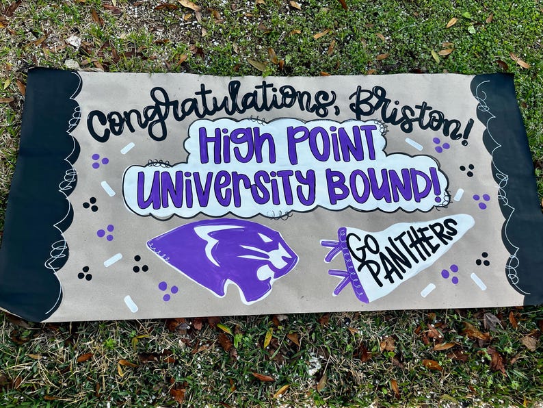 May include: A handmade banner with the text "Congratulations, Briston! High Point University Bound!" in black and purple. It features a purple panther logo and a white pennant that says "Go Panthers". The banner has black borders and is on a tan background.