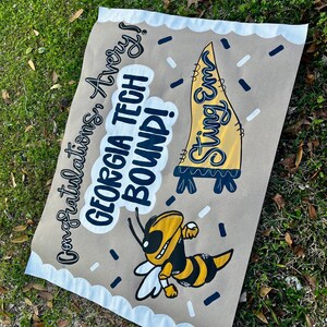 May include: A brown paper sign with the text "Congratulations Avery! GEORGIA TECH BOUND!" and a cartoon of a yellow and black bee. The sign also features a yellow pennant with the words "Sting Em".