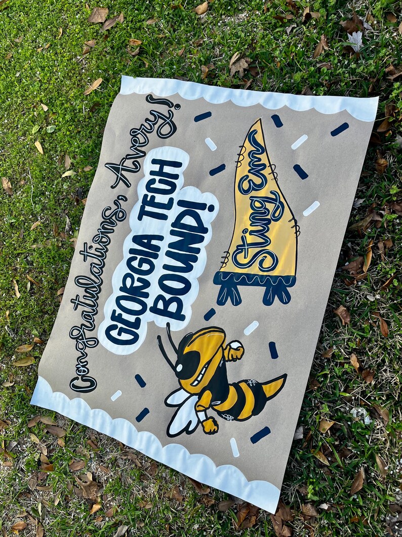 May include: A brown paper sign with the text "Congratulations Avery! GEORGIA TECH BOUND!" and a cartoon of a yellow and black bee. The sign also features a yellow pennant with the words "Sting Em".