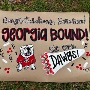 May include: A brown paper sign with the words "Congratulations, Karoline!" and "GEORGIA BOUND!" in black and red lettering. A bulldog in a red shirt and the phrase "Sic 'em Dawgs!" are also featured. Decorative confetti surrounds the text and images.