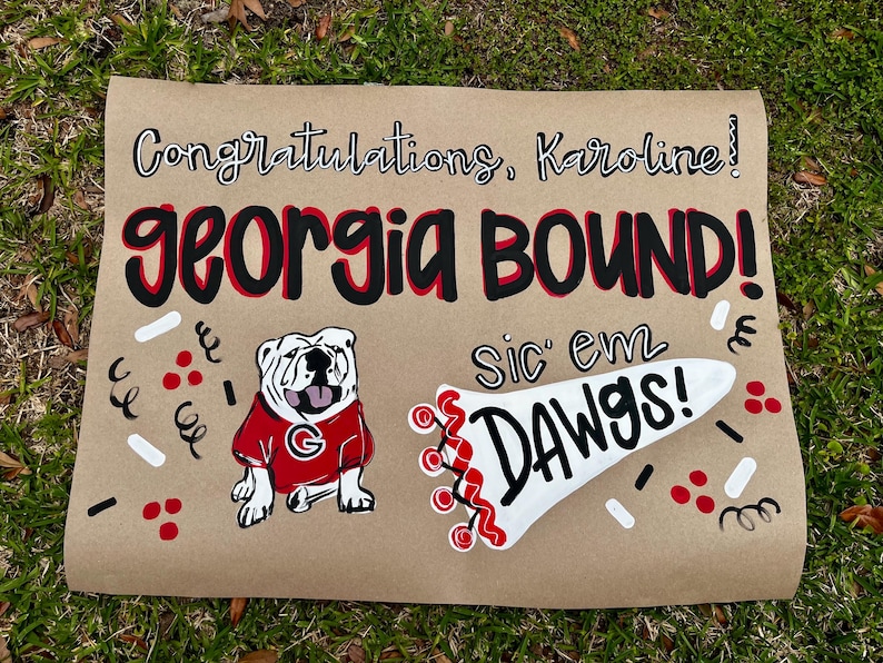 May include: A brown paper sign with the words "Congratulations, Karoline!" and "GEORGIA BOUND!" in black and red lettering. A bulldog in a red shirt and the phrase "Sic 'em Dawgs!" are also featured. Decorative confetti surrounds the text and images.