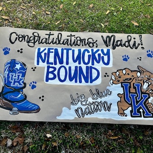 May include: A handmade banner with the words "Congratulations, Madi!" and "Kentucky Bound" in blue and black. The banner features a blue cowboy boot with a stethoscope, paw prints, and a cartoon wildcat with the letters "UK".