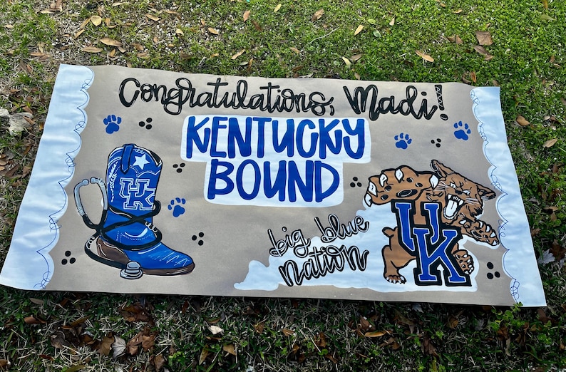 May include: A handmade banner with the text "Congratulations, Madi! KENTUCKY BOUND" in blue and black. It features a blue cowboy boot with a stethoscope, paw prints, and a UK Wildcats graphic. The words "big blue nation" are also included.