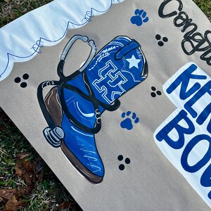 May include: A painted sign featuring a blue cowboy boot with a white star and the letters "UK" in white. A stethoscope is draped over the boot. The sign also includes the words "Congrats" and "Kit Bo" in blue and black.