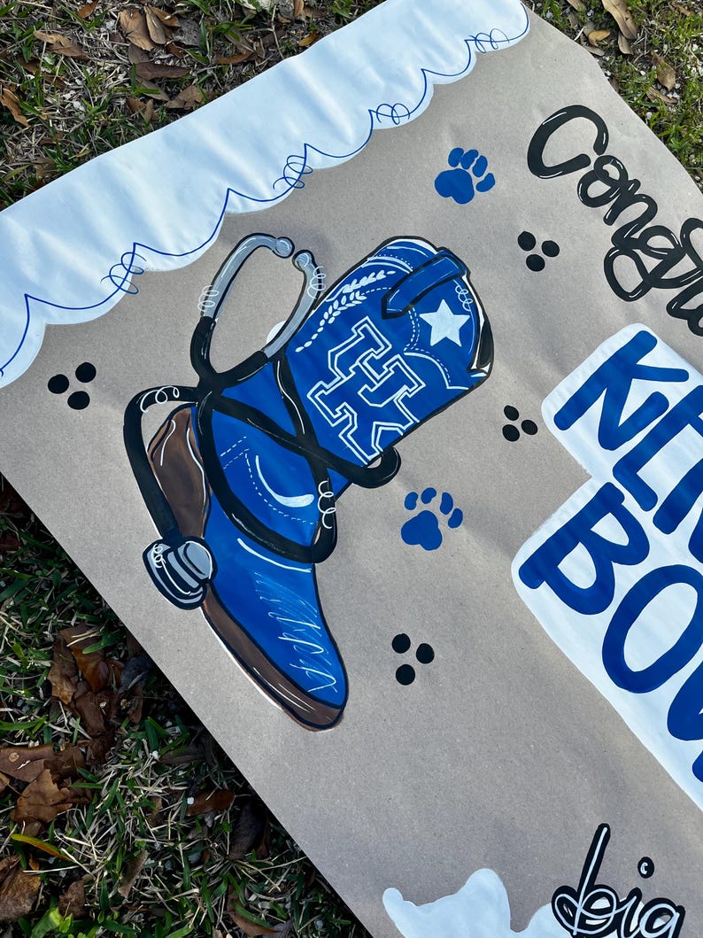 May include: A painted sign featuring a blue cowboy boot with a white star and the letters "UK" in white. A stethoscope is draped over the boot. The sign also includes the words "Congrats" and "Kit Bo" in blue and black.