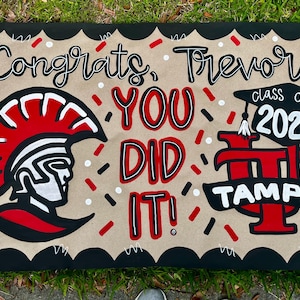 May include: A hand-painted graduation sign on a tan background with black scalloped edges. The sign says "Congrats, Trevor! You Did It!" with a Spartan helmet graphic and "class of 2026" with the word "TAMPA". Red, black, and white accents.
