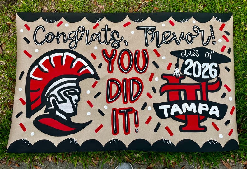 May include: A hand-painted graduation sign on a tan background with black scalloped edges. The sign says "Congrats, Trevor! You Did It!" with a Spartan helmet graphic and "class of 2026" with the word "TAMPA". Red, black, and white accents.