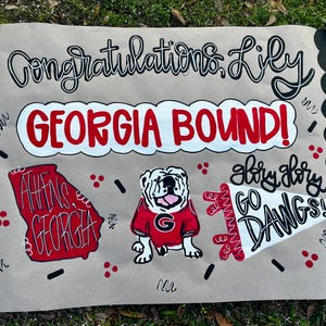 May include: A hand-painted sign on tan paper with black scalloped edges. The sign reads "Congratulations Lily GEORGIA BOUND!" with a red and white outline of Georgia, a bulldog, and "Glory Glory GO DAWGS!"