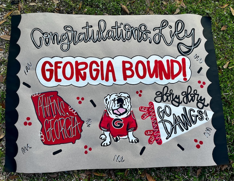 May include: A hand-painted sign on tan paper with black scalloped edges. The sign reads "Congratulations Lily GEORGIA BOUND!" with a red and white outline of Georgia, a bulldog, and "Glory Glory GO DAWGS!"