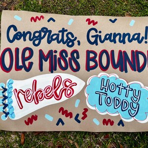 May include: A brown paper sign with the words "Congrats, Gianna! OLE MISS BOUND!" in blue and red lettering. The sign also features the word "rebels" in red and white, and "Hotty Toddy" in a blue cloud.